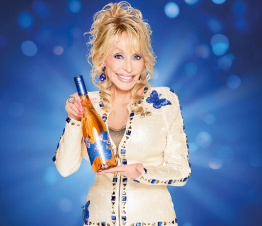 It’s Wine to Five as Dolly Parton launches new wines in UK Dolly Parton holds a bottle of Dolly Wine Rose against a blue background.