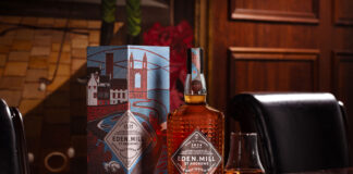 A bottle of Eden Mill Art of St Andrews whisky sits next to a nosing glass and the outer packaging for the dram on top of a wooden table.