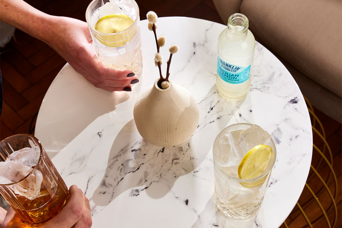 Drinks sit on a white marble table with a bottle of Franklin & Sons Sicillian Lemon on the table next to a vase.