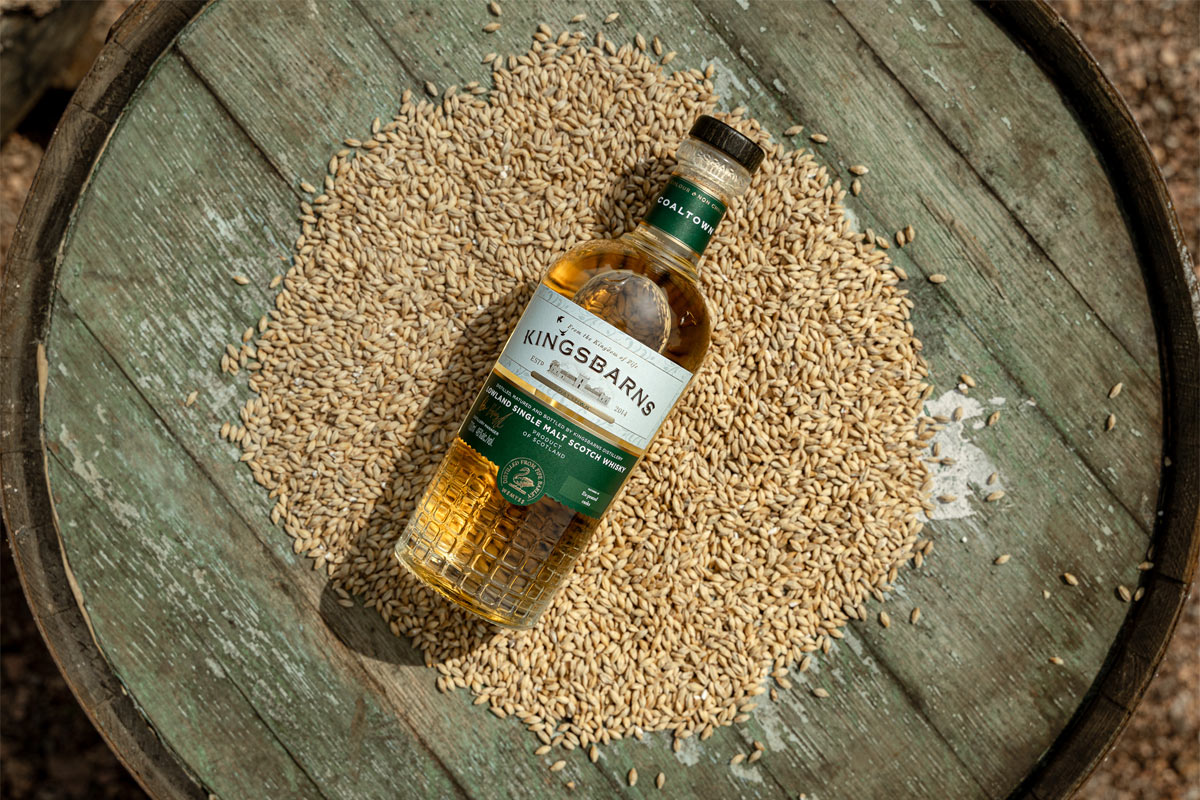 A bottle of Kingsbarns Coaltown lies on top of a pile of grain on top of a wooden barrel.