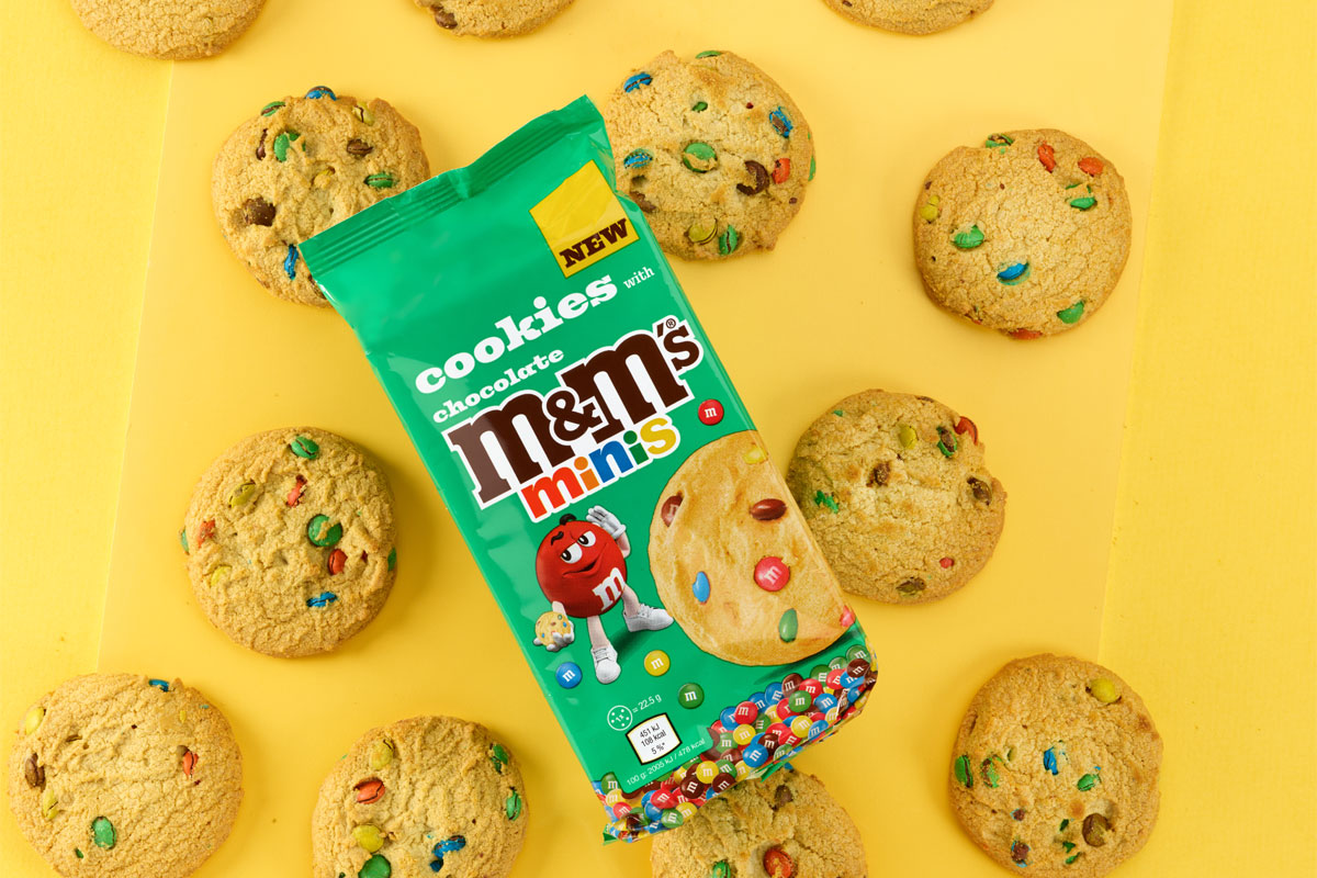 A pack of M&M's Minis Cookies sit on top of a yellow background with the cookies around the pack.