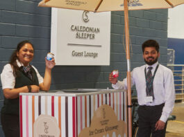 All aboard for new Mackie’s competition Two people hold small tubs of Mackie's of Scotland ice cream next to a Caledonian Sleeper branded cart with the Caledonian Sleeper sign in the background.