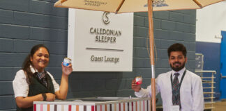 Two people hold small tubs of Mackie's of Scotland ice cream next to a Caledonian Sleeper branded cart with the Caledonian Sleeper sign in the background.