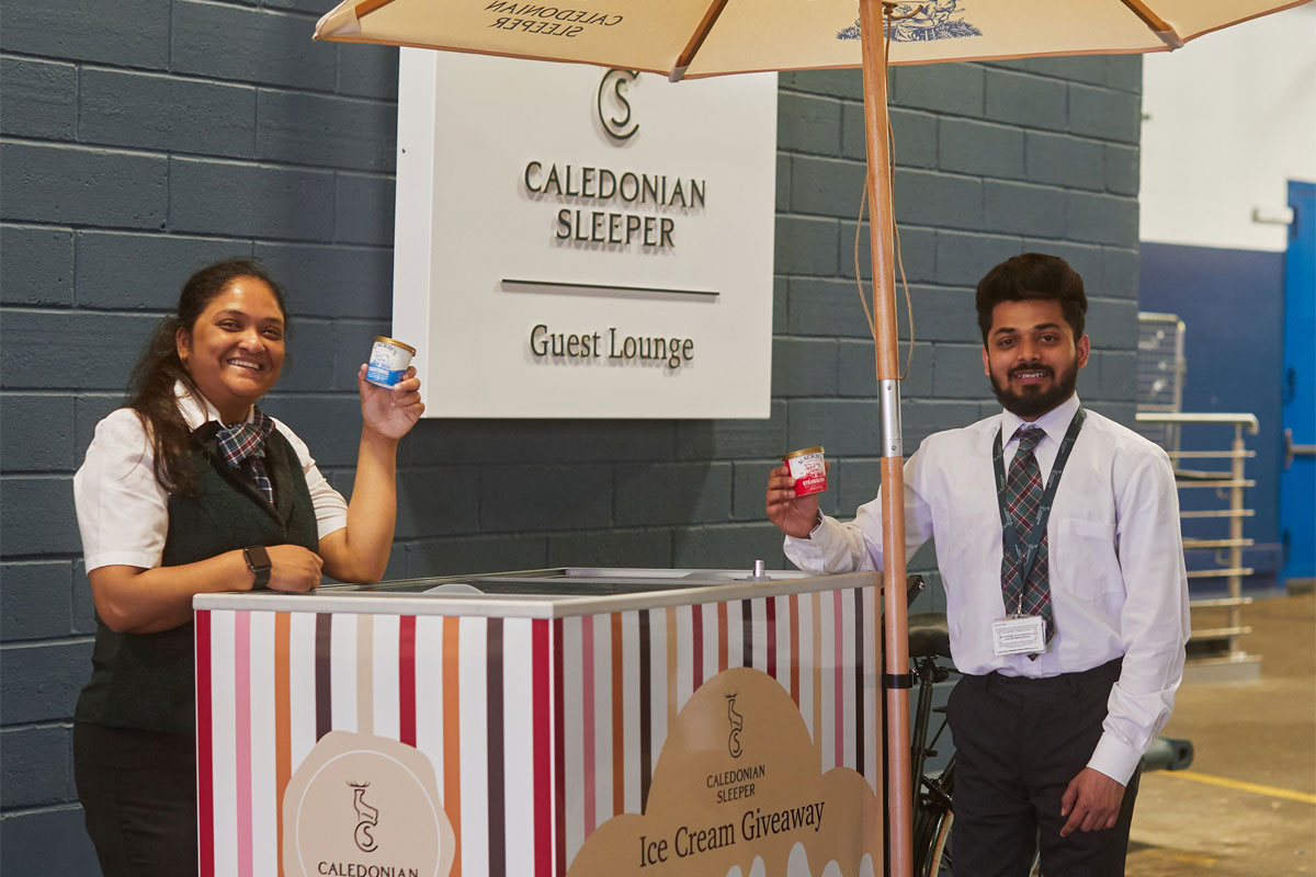 Two people hold small tubs of Mackie's of Scotland ice cream next to a Caledonian Sleeper branded cart with the Caledonian Sleeper sign in the background.