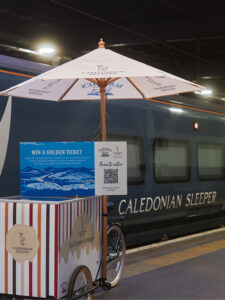 A Caledonian Sleeper ice cream cart sits in front of the Caledonian Sleeper train with scannable 'Golden Ticket' sign.