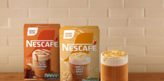 Boxes of Nescafé Dessert Edition Vanilla Cookie Dough and Sticky Toffee Pudding coffee sit on a countertop with a white tile wall behind them and a glass of coffee with a cookie next to the packs.