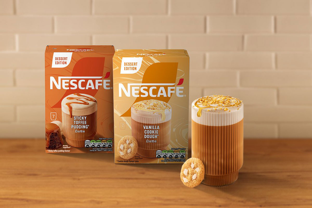 Boxes of Nescafé Dessert Edition Vanilla Cookie Dough and Sticky Toffee Pudding coffee sit on a countertop with a white tile wall behind them and a glass of coffee with a cookie next to the packs.