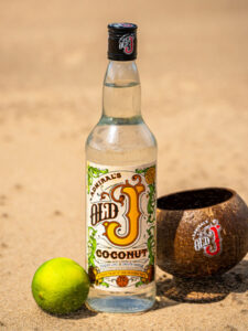 A bottle of Old J Coconut Rum stands on a beach with a coconut cup and a lime.