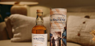 A bottle of The Balvenie A Collection of Curious Casks 14 Year Old American Bourbon Barrel stands next to its case on top of a white couch.