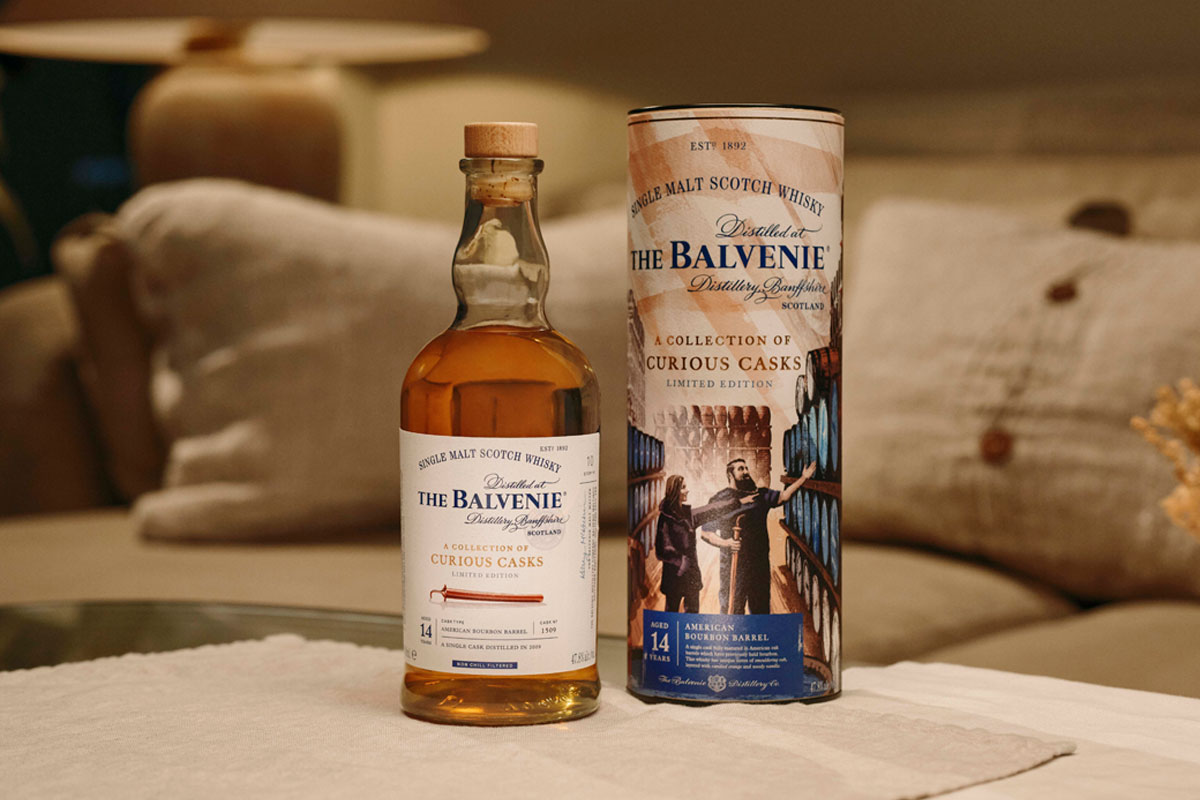 A bottle of The Balvenie A Collection of Curious Casks 14 Year Old American Bourbon Barrel stands next to its case on top of a white couch.