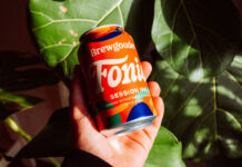 A person holds a can of Brewgooder Fairtrade Fonio grain beer in front of green leaves.