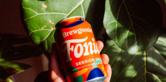 A person holds a can of Brewgooder Fairtrade Fonio grain beer in front of green leaves.