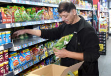 A man is stocking shelves with crisps in a convenience store.