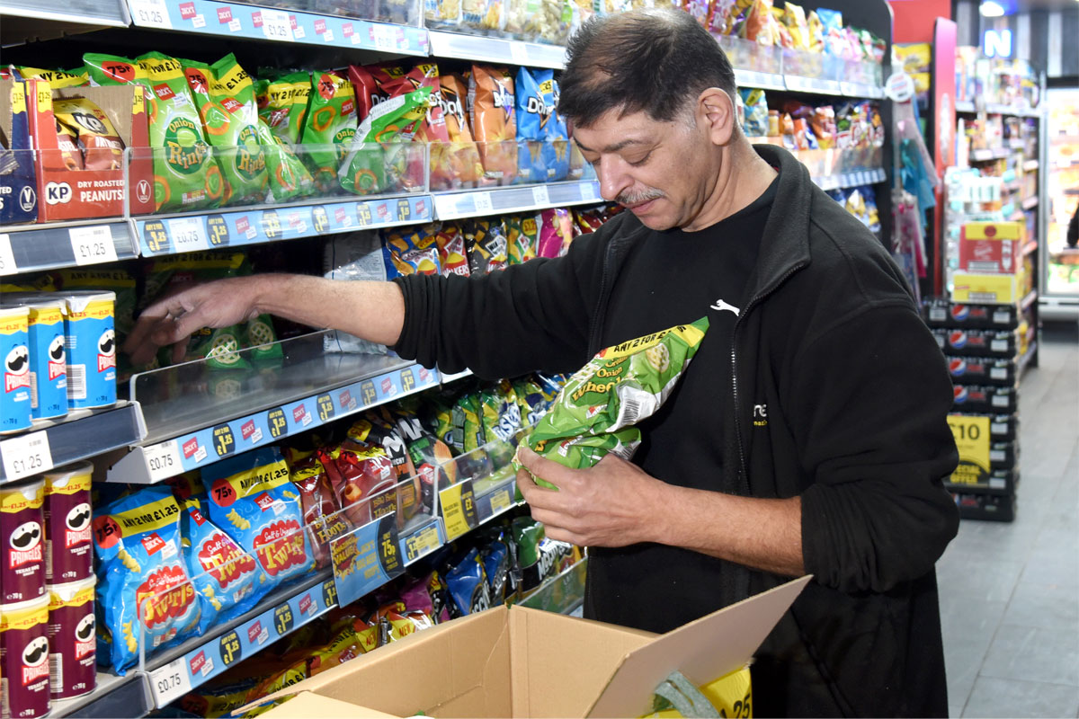 A man is stocking shelves with crisps in a convenience store.