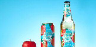 A bottle of Thatchers Juicy Cider Juicy Apple is next to a can of Thatchers Cider Juicy Apple with a red apple next to the can against a blue background with a shiny red table underneath.