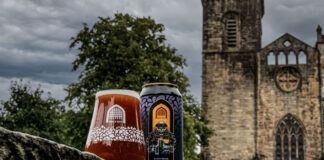 A can of Vault City Black Grape Buckie V.2 stands next to a glass filled with the sour beer in front of the ruins of a church building.