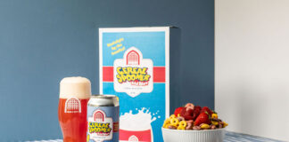 A can of Vault City Cereal Spooner sour beer stands in front of a cereal box advertising the sour beer with it poured out in a Vault City branded glass and a bowl of cereal and fruit to the right.