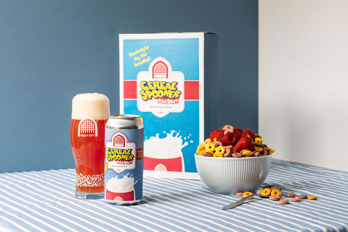 A can of Vault City Cereal Spooner sour beer stands in front of a cereal box advertising the sour beer with it poured out in a Vault City branded glass and a bowl of cereal and fruit to the right.