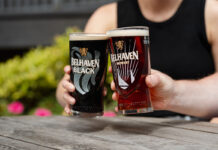 Two people sit holding Belhaven branded glasses with beer inside them and clink them together.