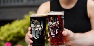 Two people sit holding Belhaven branded glasses with beer inside them and clink them together.