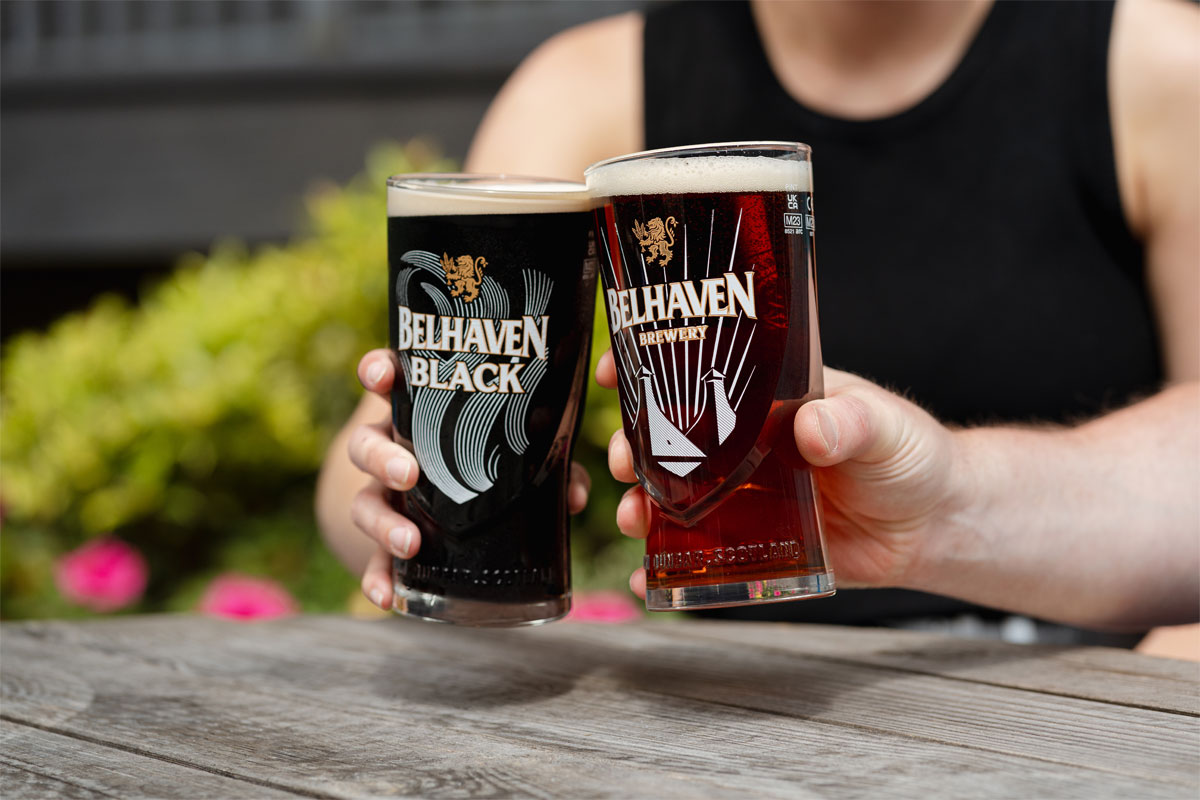 Two people sit holding Belhaven branded glasses with beer inside them and clink them together.