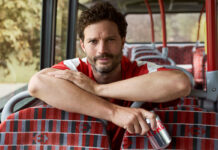 Jamie Dornan sits on a bus leaning over seats holding a can of Diet Coke.