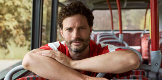 Jamie Dornan sits on a bus leaning over seats holding a can of Diet Coke.