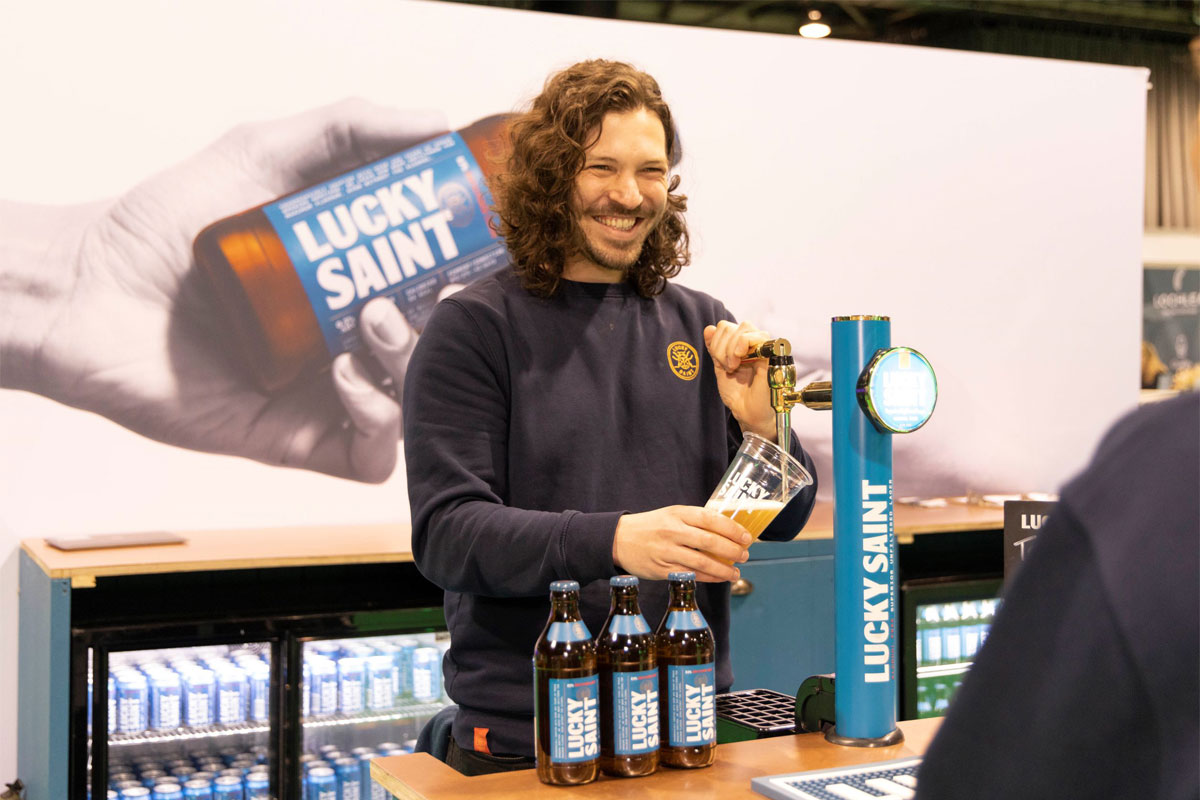 A person stands at a Lucky Saint branded beer tap pulling a pint with bottle of Lucky Saint in front of him.