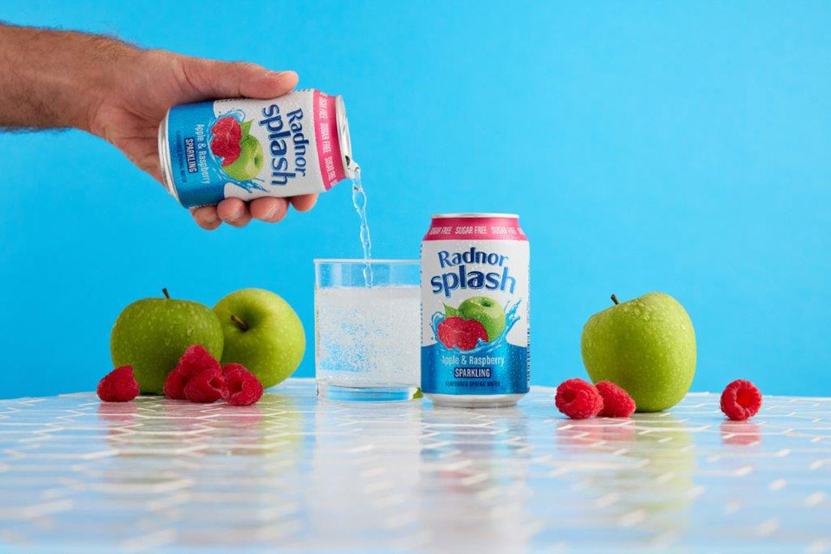 Lifestyle image of a person pouring a can of Radnor Splash into a glass with another can next to the glass and fruit around it.