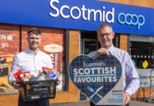 Two people stand outside a Scotmid store, one person holds a sign for the Scotmid Scottish Favourites Competition while the other holds a basket filled with Scottish produce.