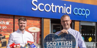 Two people stand outside a Scotmid store, one person holds a sign for the Scotmid Scottish Favourites Competition while the other holds a basket filled with Scottish produce.