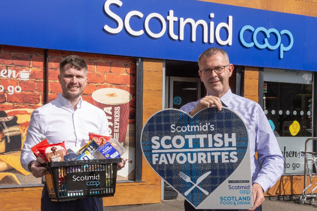 Two people stand outside a Scotmid store, one person holds a sign for the Scotmid Scottish Favourites Competition while the other holds a basket filled with Scottish produce.