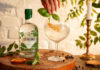 A bottle of Stirling Distillery's Classic Gin stands next to a glass with a drink serve as a hand places some basil into the drink.