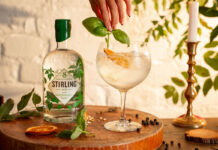 A bottle of Stirling Distillery's Classic Gin stands next to a glass with a drink serve as a hand places some basil into the drink.