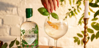 A bottle of Stirling Distillery's Classic Gin stands next to a glass with a drink serve as a hand places some basil into the drink.