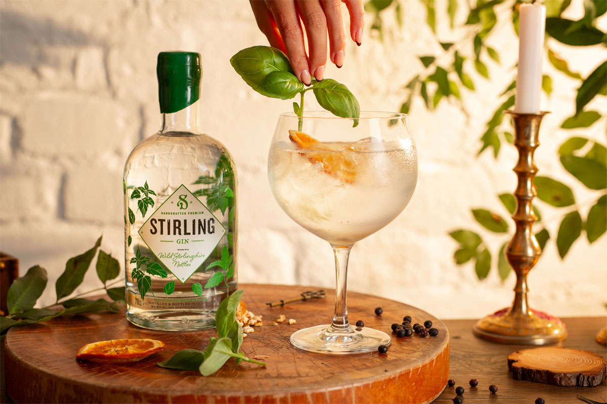 A bottle of Stirling Distillery's Classic Gin stands next to a glass with a drink serve as a hand places some basil into the drink.