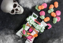 A packet of Swizzels Squashies Squeletons lie on a table with the sweets spilled out, above the packet is a skull and fog at the top and bottom of the image.