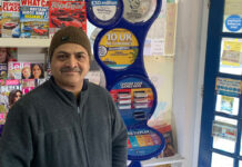 Ravindrasinh Bhatti, retailer of The Corner Shop in Hythe, stands in front of a National Lottery game station with magazine behind him.