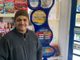 Allwyn awards £40k to 22 retailers Ravindrasinh Bhatti, retailer of The Corner Shop in Hythe, stands in front of a National Lottery game station with magazine behind him.