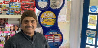 Ravindrasinh Bhatti, retailer of The Corner Shop in Hythe, stands in front of a National Lottery game station with magazine behind him.