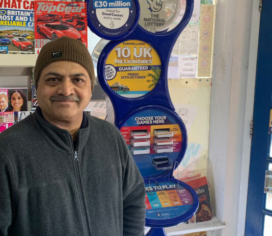 Allwyn awards £40k to 22 retailers Ravindrasinh Bhatti, retailer of The Corner Shop in Hythe, stands in front of a National Lottery game station with magazine behind him.