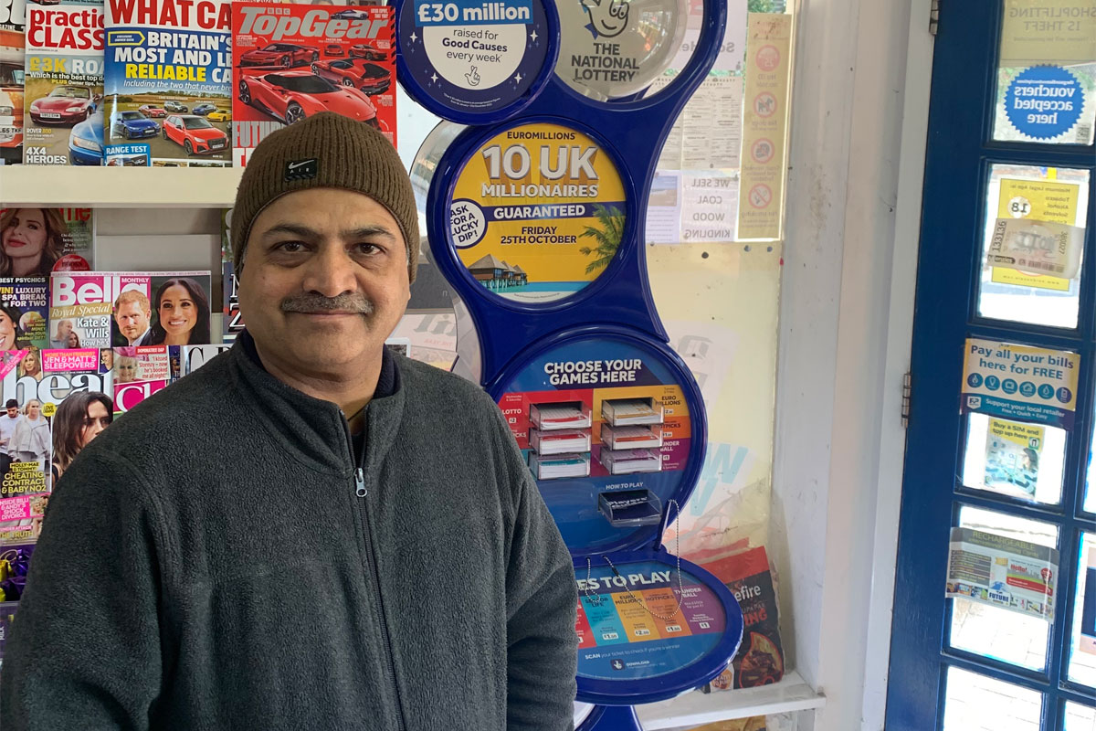 Ravindrasinh Bhatti, retailer of The Corner Shop in Hythe, stands in front of a National Lottery game station with magazine behind him.