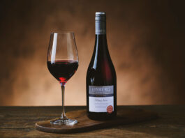 A bottle of Eisberg Pinot Noir Non-Alcoholic Wine stands next to a glass with the wine in it on a wooden table in a darkly lit room.