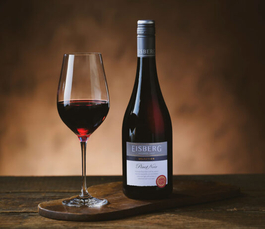 Eisberg builds non-alc wine option for Christmas A bottle of Eisberg Pinot Noir Non-Alcoholic Wine stands next to a glass with the wine in it on a wooden table in a darkly lit room.