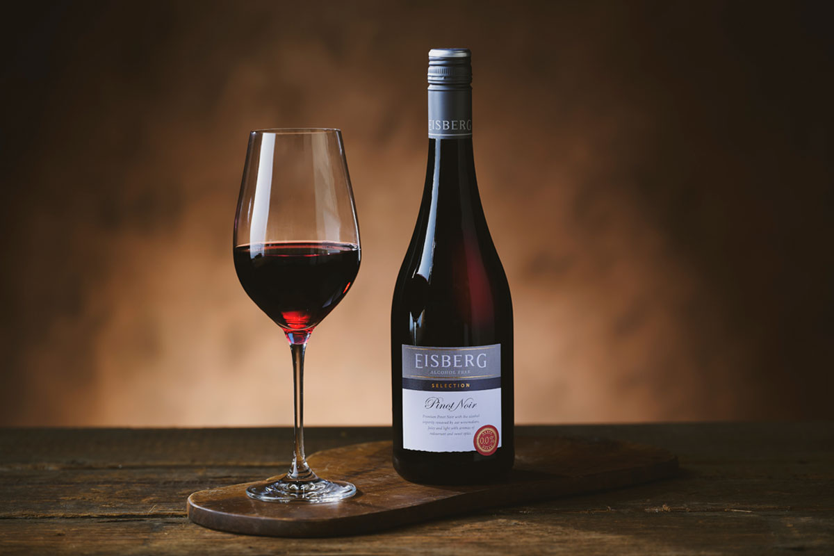 A bottle of Eisberg Pinot Noir Non-Alcoholic Wine stands next to a glass with the wine in it on a wooden table in a darkly lit room.