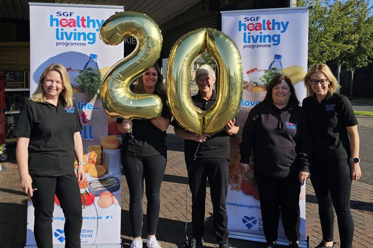 The SGF Healthy Living Programme team stand holding a two balloon and a zero balloon together in front of HLP branded stands.