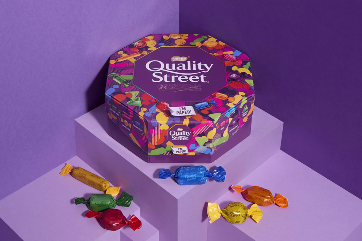 Promotional image of Quality Street paper tubs featuring the tub and sweets from the tub against a purple background.