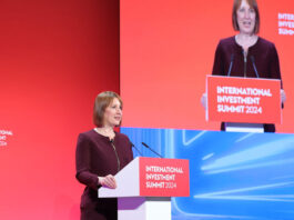 Rachel Reeves, chancellor of the exchequer, stands at a podium delivering a speech with a TV screen featuring her to her left.