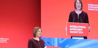 Rachel Reeves, chancellor of the exchequer, stands at a podium delivering a speech with a TV screen featuring her to her left.