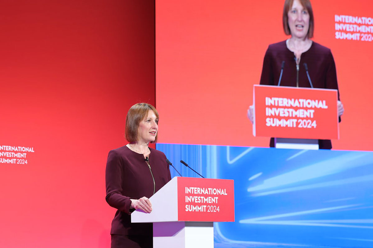 Rachel Reeves, chancellor of the exchequer, stands at a podium delivering a speech with a TV screen featuring her to her left.